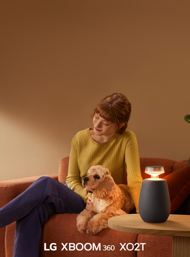 Sieviete sē? uz dīvāna ar suni, klausoties mūziku ar LG XBOOM 360 XO2T.