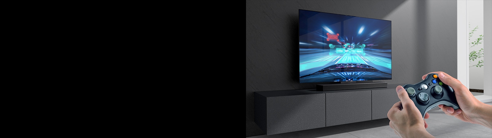 Ska?as panelis novietots uz skapī?a, pievienotajā TV ekrānā redzama auto sacīk?u spēles aina. Attēla labajā apak?ējā malā redzama spē?u konsole, kuru tur abās rokās.