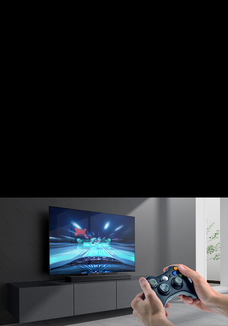 Ska?as panelis novietots uz skapī?a, pievienotajā TV ekrānā redzama auto sacīk?u spēles aina. Attēla labajā apak?ējā malā redzama spē?u konsole, kuru tur abās rokās.