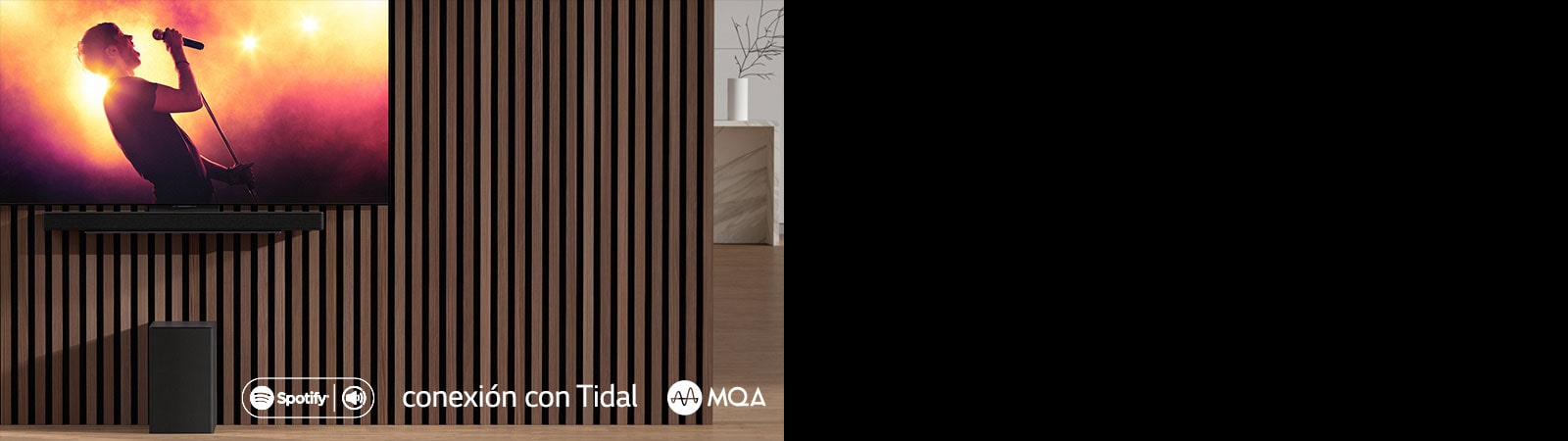 LG OLED C ir piestiprināts pie sienas, zem tā uz ekskluzīvā kron?teina ir piestiprināts LG Sound Bar SC9S. Zemo frekven?u pastiprinātājs ir novietots zem tā. TV ekrānā redzama koncerta aina.