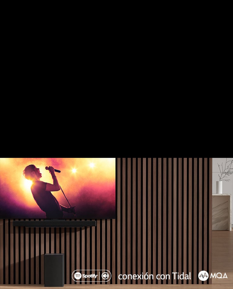 LG OLED C ir piestiprināts pie sienas, zem tā uz ekskluzīvā kron?teina ir piestiprināts LG Sound Bar SC9S. Zemo frekven?u pastiprinātājs ir novietots zem tā. TV ekrānā redzama koncerta aina.