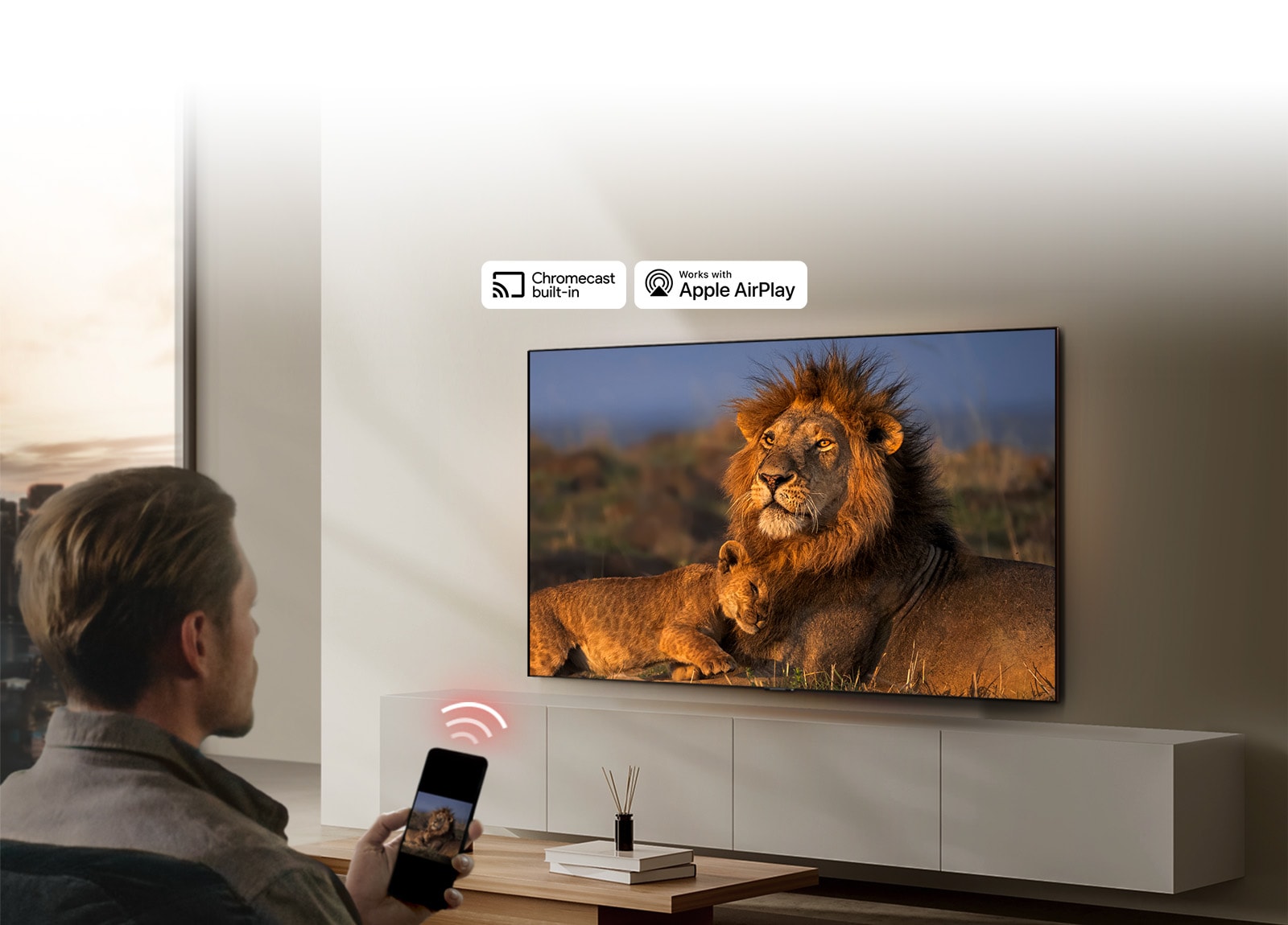 LG TV postavljen na zid u dnevnoj sobi prikazuje lava i lavi?a. ?ovek sedi u prvom planu i u ruci dr?i pametni telefon na kojem se vidi ista slika lavova. Ilustracija sa tri neonsko crvene zakrivljene trake vidi se odmah iznad pametnog telefona koji je usmeren ka TV ure?aju.