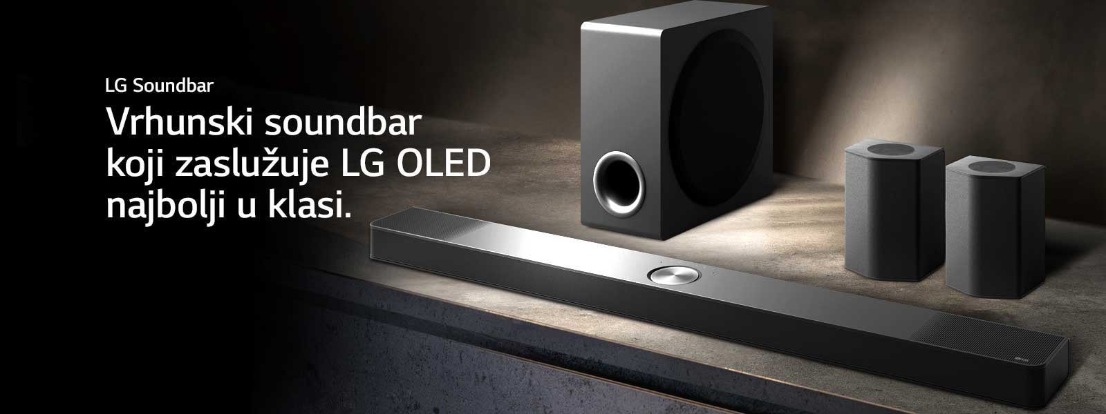 LG Soundbar, zadnji zvu?nici i niskotonac postavljeni su iskosa na sme?oj drvenoj polici u zamra?enoj crnoj prostoriji sa svetlom samo iznad zvu?nog sistema.