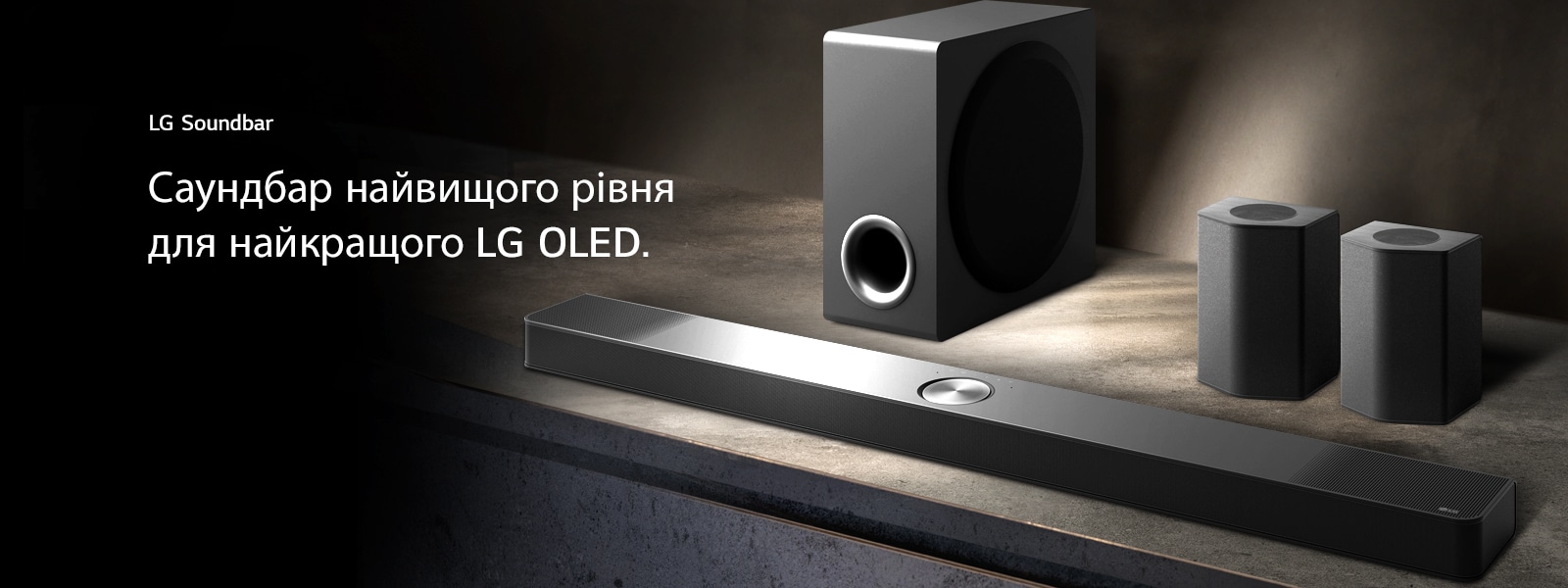 LG Soundbar, задн? динам?ки та сабвуфер (вигляд п?д кутом) на коричнев?й дерев’ян?й полиц? в чорн?й к?мнат?, огорнут?й темрявою, де св?тло пада? лише на звукову систему.